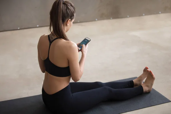 Young attractive woman sitting on yoga mat texting on phone