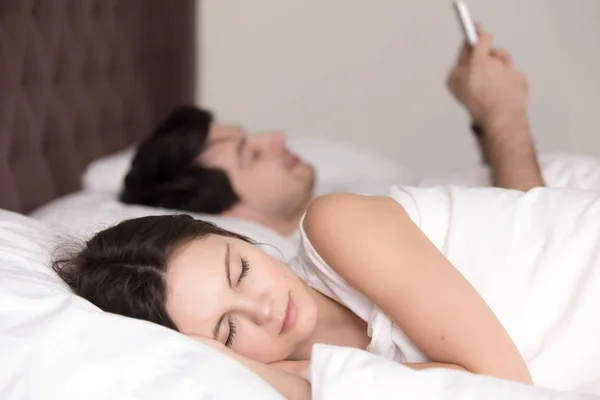 Mujer joven durmiendo mientras su novio usando el teléfono inteligente en la cama — Foto de Stock