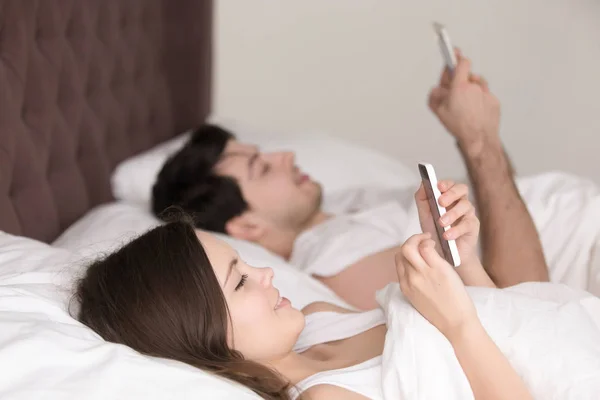 Pareja feliz cada uno usando su propio teléfono inteligente acostado en la cama — Foto de Stock