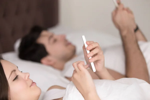 Primer plano de la pareja joven usando teléfonos inteligentes móviles acostados en la cama — Foto de Stock