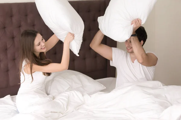 Veselá holka a chlap bojuje s polštáři sedět na posteli — Stock fotografie