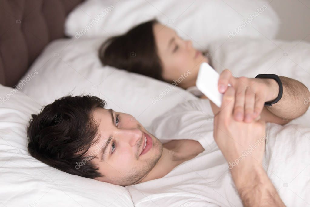 Young guy using mobile phone while girlfriend sleeping in bed