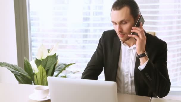 Νεαρός επιχειρηματίας, απάντηση σε κλήση ενώ χρησιμοποιείτε φορητό υπολογιστή, μιλάει στο τηλέφωνο — Αρχείο Βίντεο