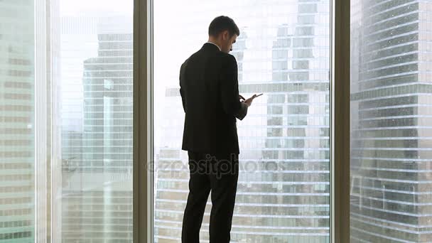 Rear view at businessman standing near full-length window using tablet — Stock Video