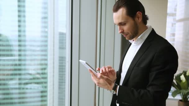 Atractivo hombre de negocios sonriente usando tableta de pie en la ventana en la oficina — Vídeos de Stock