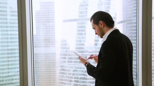 Confident happy businessman standing at office window using tablet smartphone