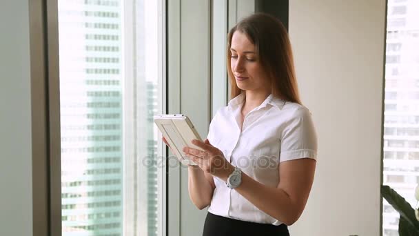 Досить молода бізнес-леді стоїть у вікні офісу за допомогою планшета — стокове відео