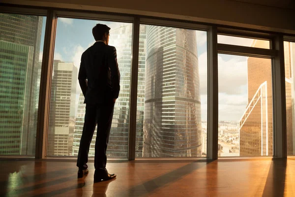 Thoughtful businessman looking out of big window at sunset city — Stock Photo, Image