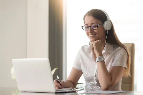 Souriant jeune femme d'affaires portant des écouteurs regardant PC lap — Photo