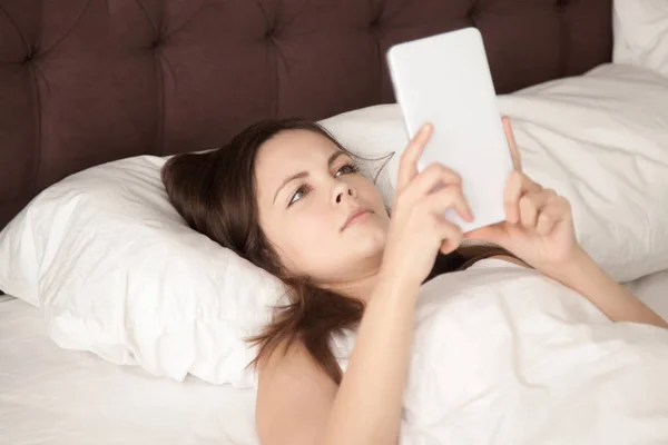 Young woman holding digital tablet lying in bed, reading e-book — Stock Photo, Image