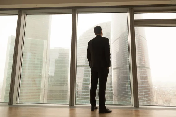 Businessman standing near full-length window looking at city, ba — Stock Photo, Image