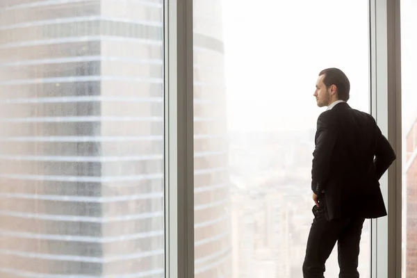 Serious young businessman standing in modern office, looking out — Stock Photo, Image