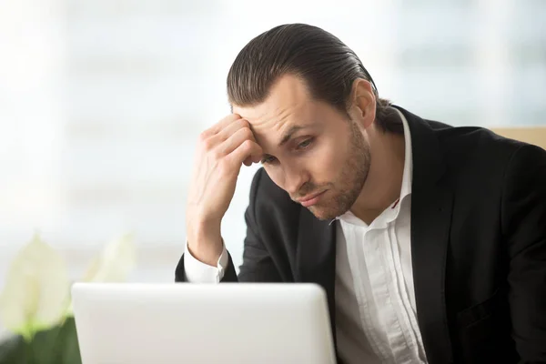 Puzzled young businessman looking at laptop screen at workplace.