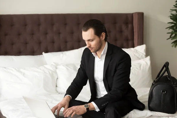Mladý podnikatel, sedí na posteli, pracovat na notebooku v hotelu. — Stock fotografie