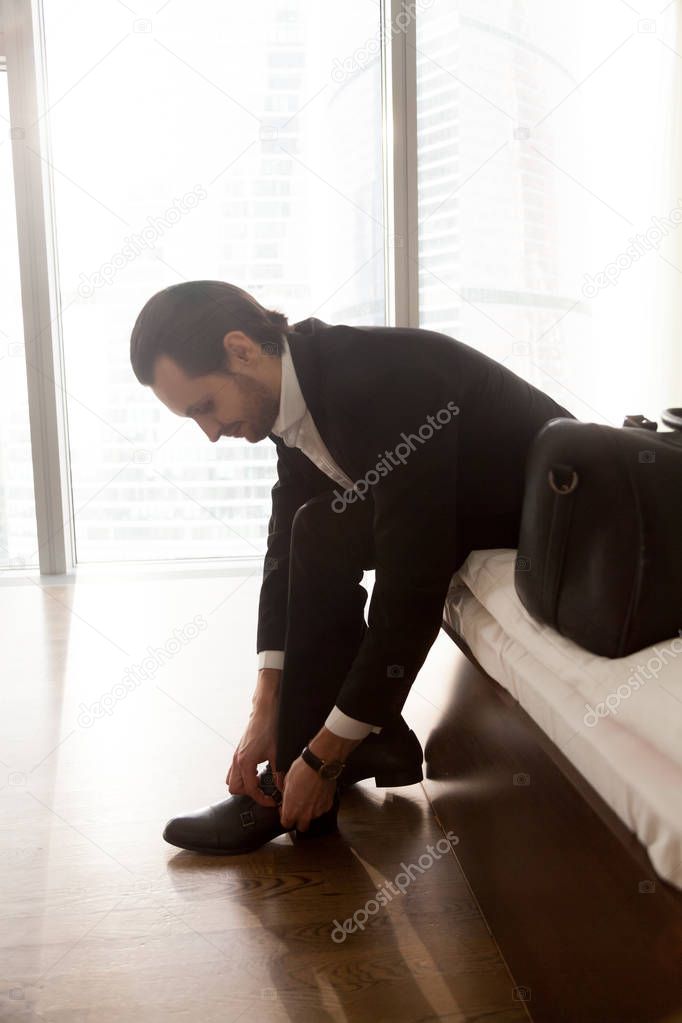 Businessman sitting on bed, putting shoes on in hotel room.