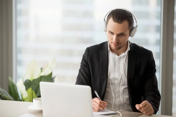 Empresario en auriculares tomando notas delante de la computadora portátil . — Foto de Stock