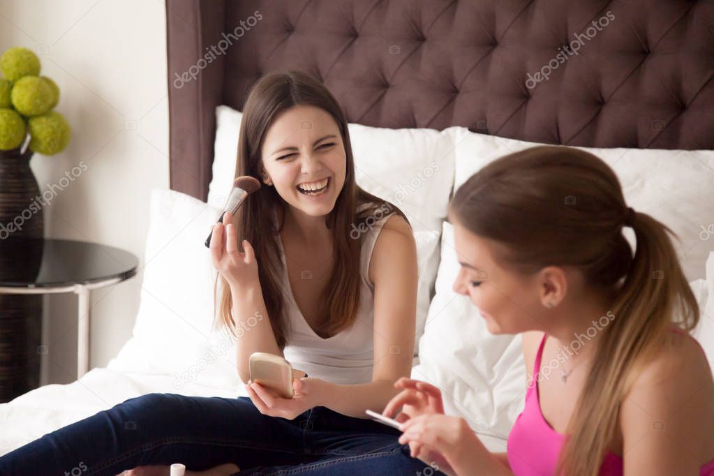 Two young girlfriends doing makeup and chatting in bedroom.