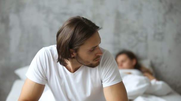 Задумчивый обеспокоенный мужчина сидит на кровати, спящая женщина на заднем плане — стоковое видео
