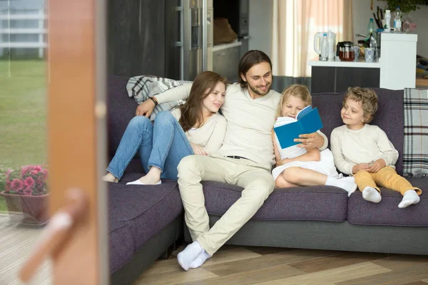 Сім'я з дітьми читає книгу, сидячи на дивані вдома — стокове фото