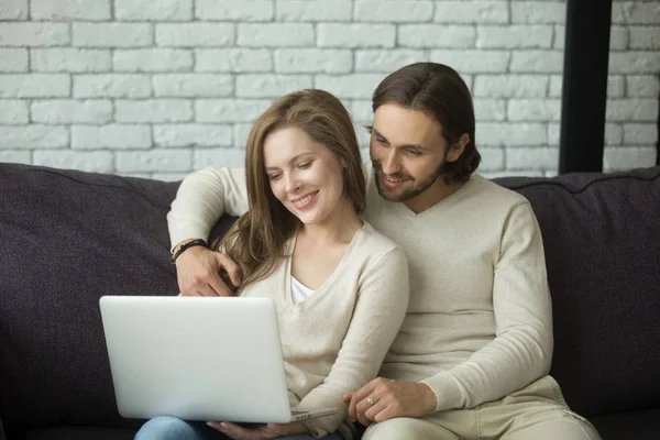 Молода пара сидить на дивані, обіймаючись ноутбуком вдома — стокове фото