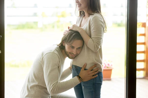 Счастливый мужчина прислушивается к беременной жене живота дома — стоковое фото