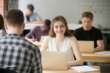 Paylaşılan Office, işkadını alan coworking içinde dizüstü bilgisayar ile birlikte çalışma, Genç freelancer öğrenci stajyer çalışan masasında oturan kamera bakarak gülümseyen çekici kadın portresi