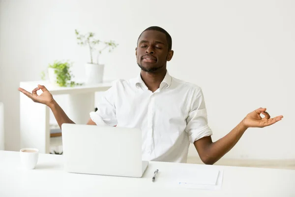 Calm happy african man meditating at office desk with laptop