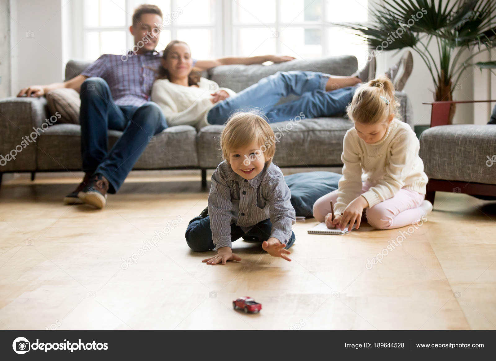 Kids Playing On Floor Pas Relaxing Sofa At Home Stock Cute Kids Playing While Pas Relaxing Sofa At Home Together Smiling Active Boy Entertaining With Toy Car Near His Sister On Floor Happy Family Spending
