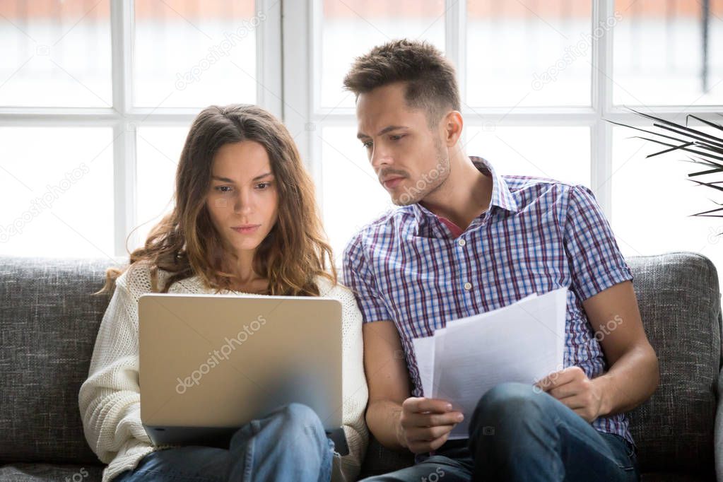 Focused worried couple paying bills online on laptop with docume