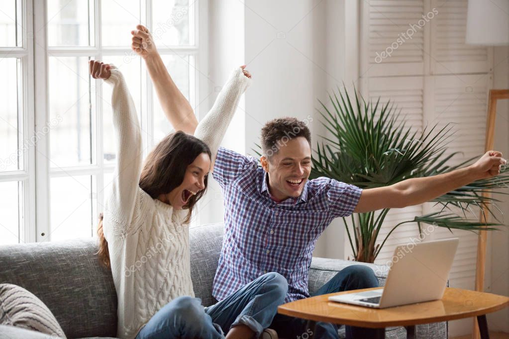 Excited couple raising hands looking at laptop celebrating onlin