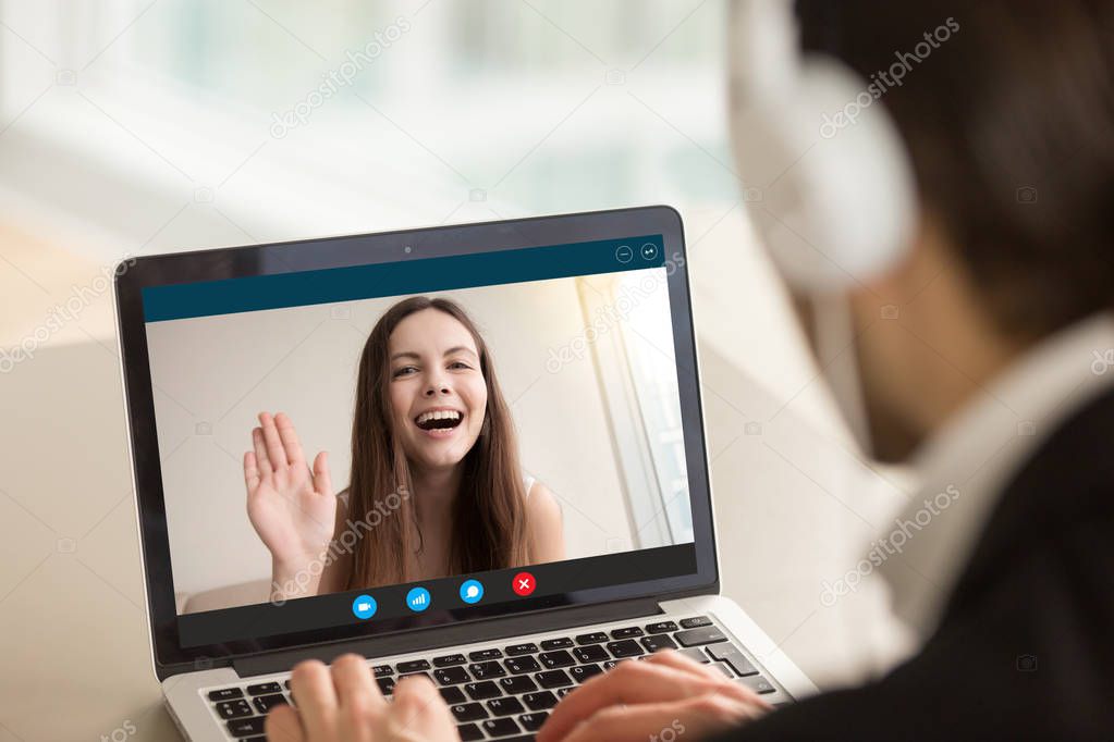 Young girl waving from laptop screen at man in headphones.