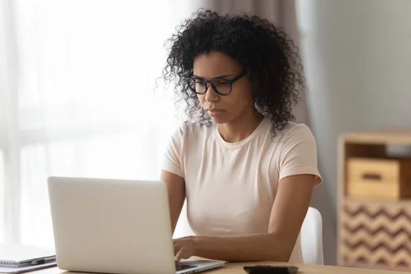 Сосредоточенная черная женщина в очках, работающая за ноутбуком — стоковое фото
