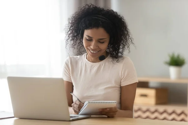 Uśmiechnięta birasowa kobieta robi notatki studiując na laptopie w domu — Zdjęcie stockowe