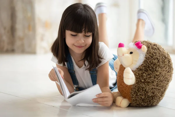 Adorable girl laying on warm floor with favorite toy, reading. — Stock Photo, Image