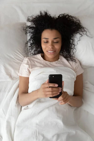 Chica negra usando celular despertando en la cama por la mañana — Foto de Stock