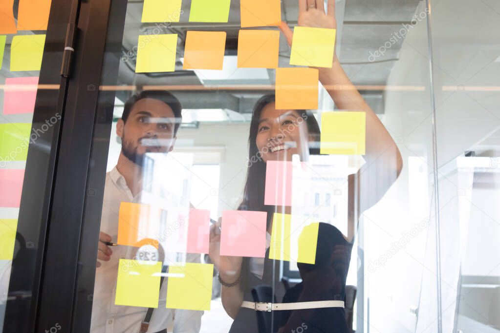 Smiling diverse employees looking at sticky papers on glass wall