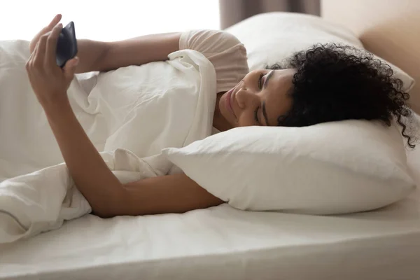 Mulher afro-americana feliz usando telefone, deitado na cama — Fotografia de Stock