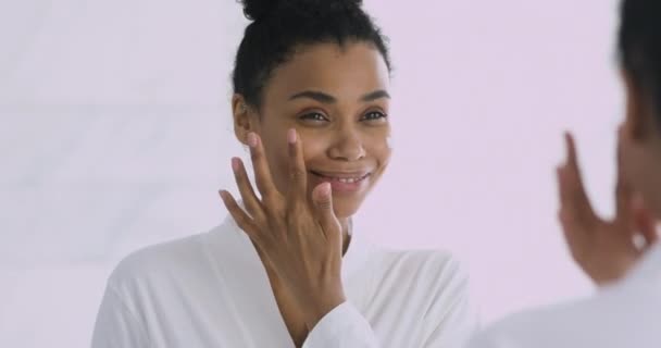 Щаслива молода африканська жінка, що застосовує крем для обличчя, дивиться у дзеркало — стокове відео