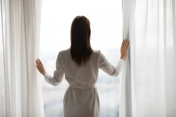 Jonge vrouw open gordijn naar buiten kijkend raam, achteruitkijkspiegel — Stockfoto