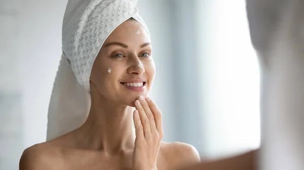 Sorridente bella signora applicare crema per la pelle guardare nello specchio — Foto Stock