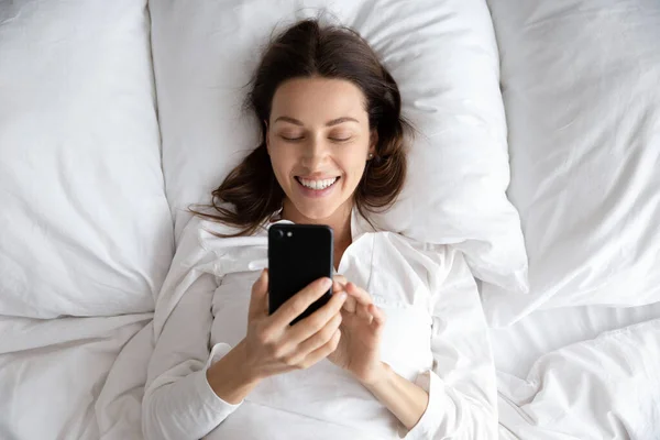 Mujer joven feliz usando el teléfono acostado en la cama por la mañana — Foto de Stock