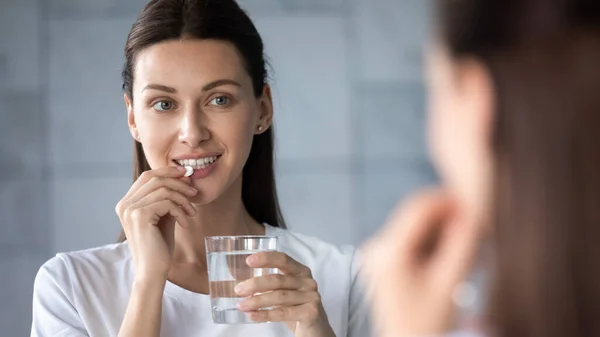 Jong gezond vrouw holding pil water glas kijken in spiegel — Stockfoto