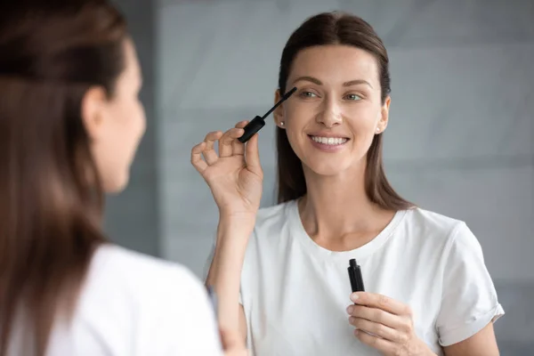 Glückliche attraktive Frau, die Mascara auf Wimpern aufträgt und in den Spiegel schaut — Stockfoto