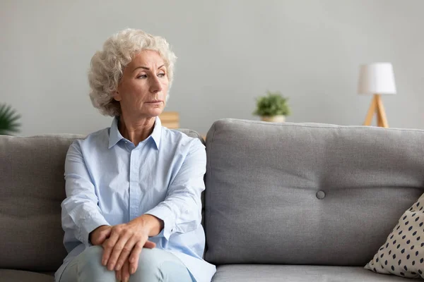 Zamyślona starsza babcia patrząc daleko myśląc o samotności na kanapie — Zdjęcie stockowe