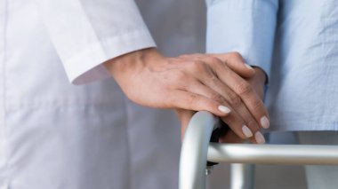 Yaşlı bir hastanın elini tutan kadın doktor yürüyüş çerçevesi kullanıyor, yakın plan.