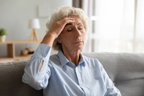 Verärgerte Seniorin mit Schwindelgefühlen sitzt mit Kopfschmerzen auf Sofa — Stockfoto