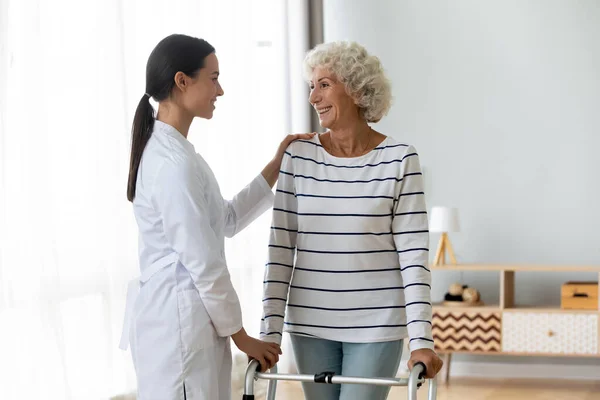 Ung kvinnelig fysioterapeut hjelper skadet gammel bestemor med gåstol – stockfoto