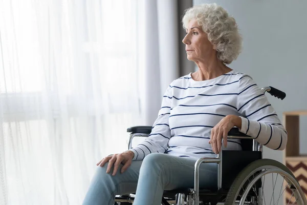 Triste mulher idosa paraplégica sentar-se em cadeira de rodas olhar através da janela — Fotografia de Stock