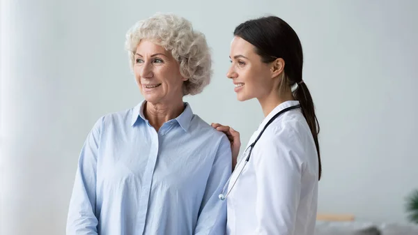 Feliz abuela vieja y joven enfermera cuidadora mirando hacia otro lado — Foto de Stock