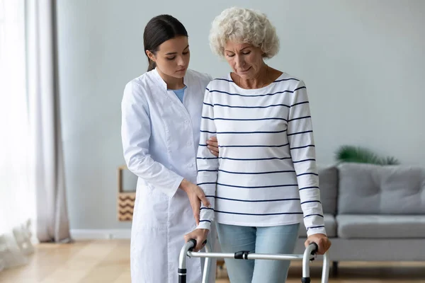 Young woman caregiver helping disabled old grandma patient using walker — Stock Photo, Image